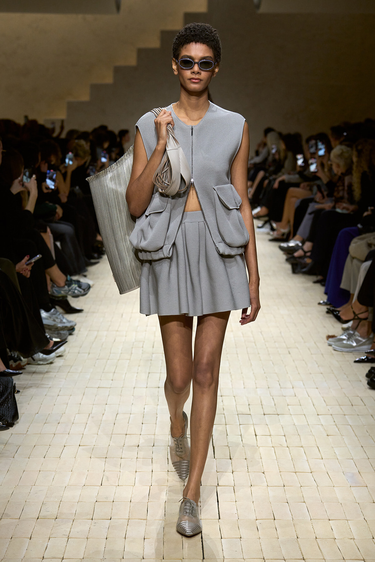 Model walks the runway in a light gray sleeveless utility dress with large front pockets, carrying a rolled bag and wearing sunglasses.