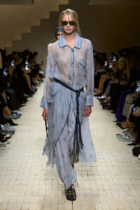 Runway model walks the catwalk in a sheer pale blue dress with a dark rope belt and sunglasses, audience watching