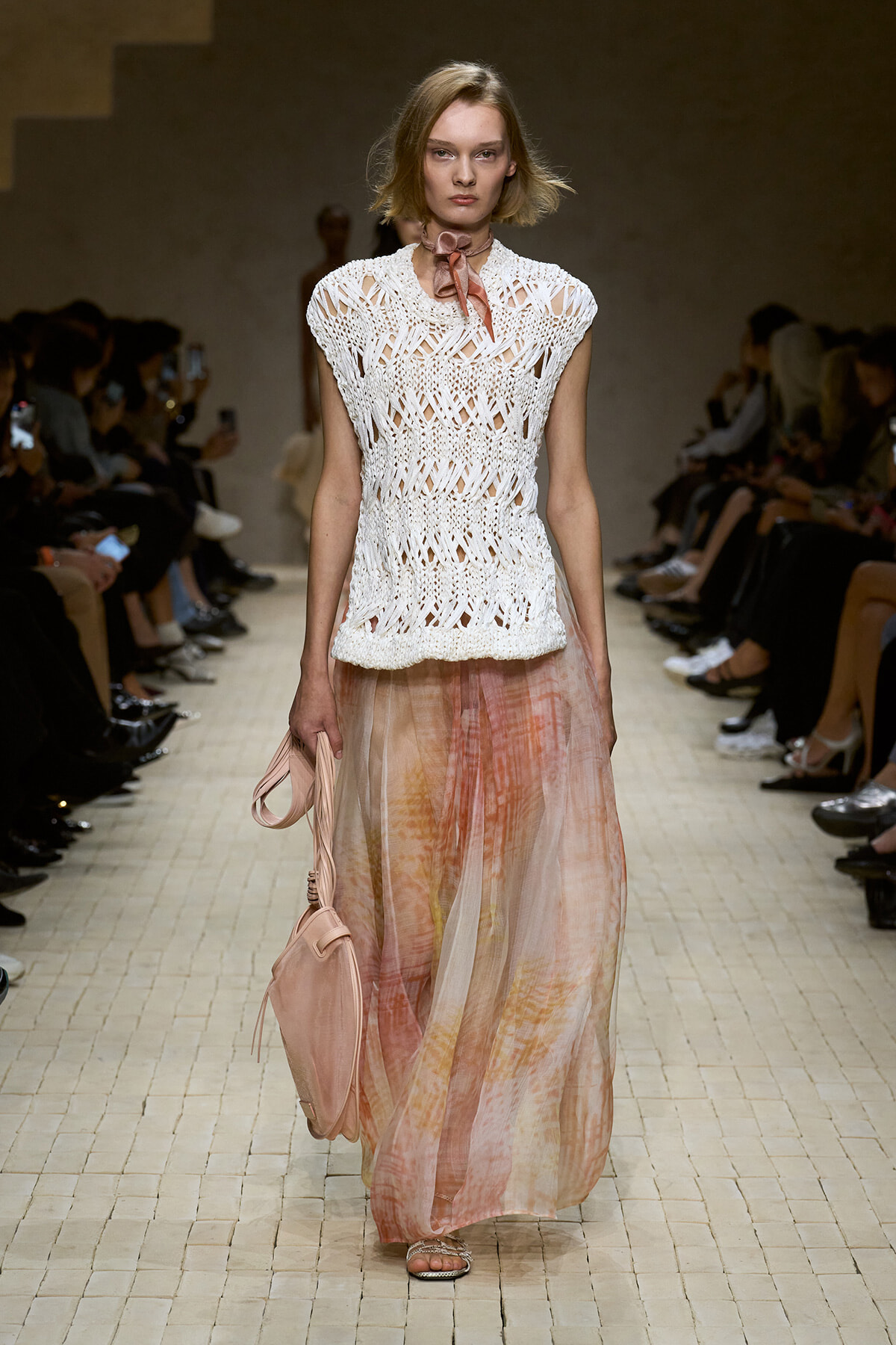 Model walks the runway in a white crochet sleeveless top and long pastel pink tie-dye skirt, holding a pink bag.