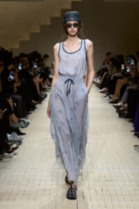Model walks the runway in a light blue, sleeveless maxi dress with a drawstring waist and blue sandals, wearing dark sunglasses and a textured cap—audience along the catwalk