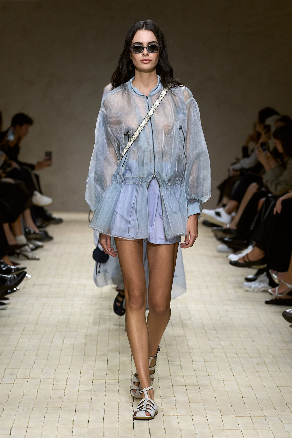 Model walks a fashion show wearing a translucent blue windbreaker over light shorts, with a white crossbody bag and small sunglasses.