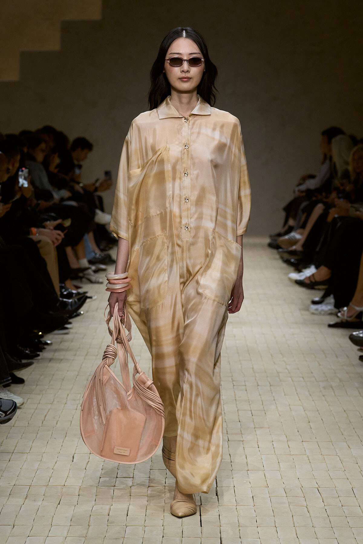 Model walks the runway in a light beige satin jumpsuit with a pink handbag and pink sunglasses.