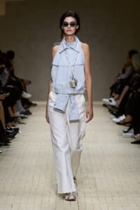 Model on a runway wearing a light blue sleeveless top with a high collar and waist tie, paired with white wide-leg pants and sunglasses.