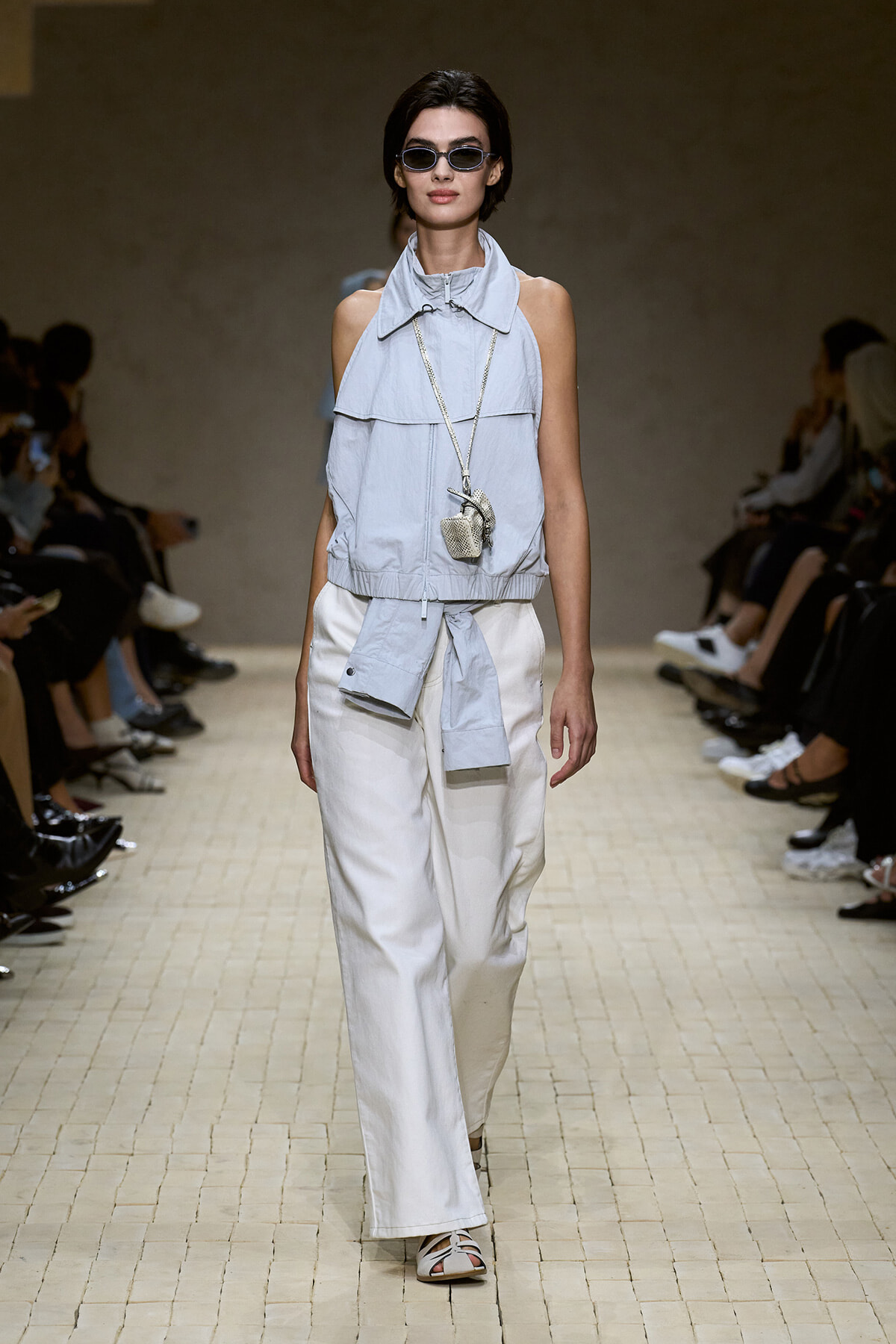 Model on a runway wearing a light blue sleeveless top with a high collar and waist tie, paired with white wide-leg pants and sunglasses.