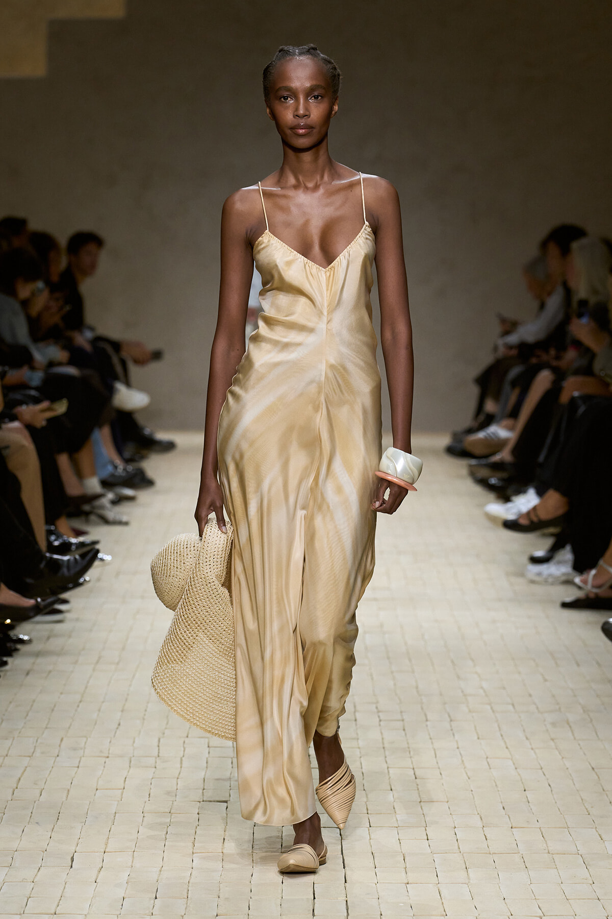 Model walking a runway wearing a silky champagne slip dress with spaghetti straps, carrying a woven tote and wearing matching beige flats and a chunky bracelet.