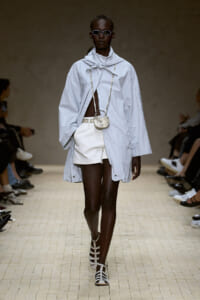 Model walks the runway in a pale oversized cape-like light coat, white shorts, and strappy sandals, accessorized with a tiny crossbody bag and sunglasses.