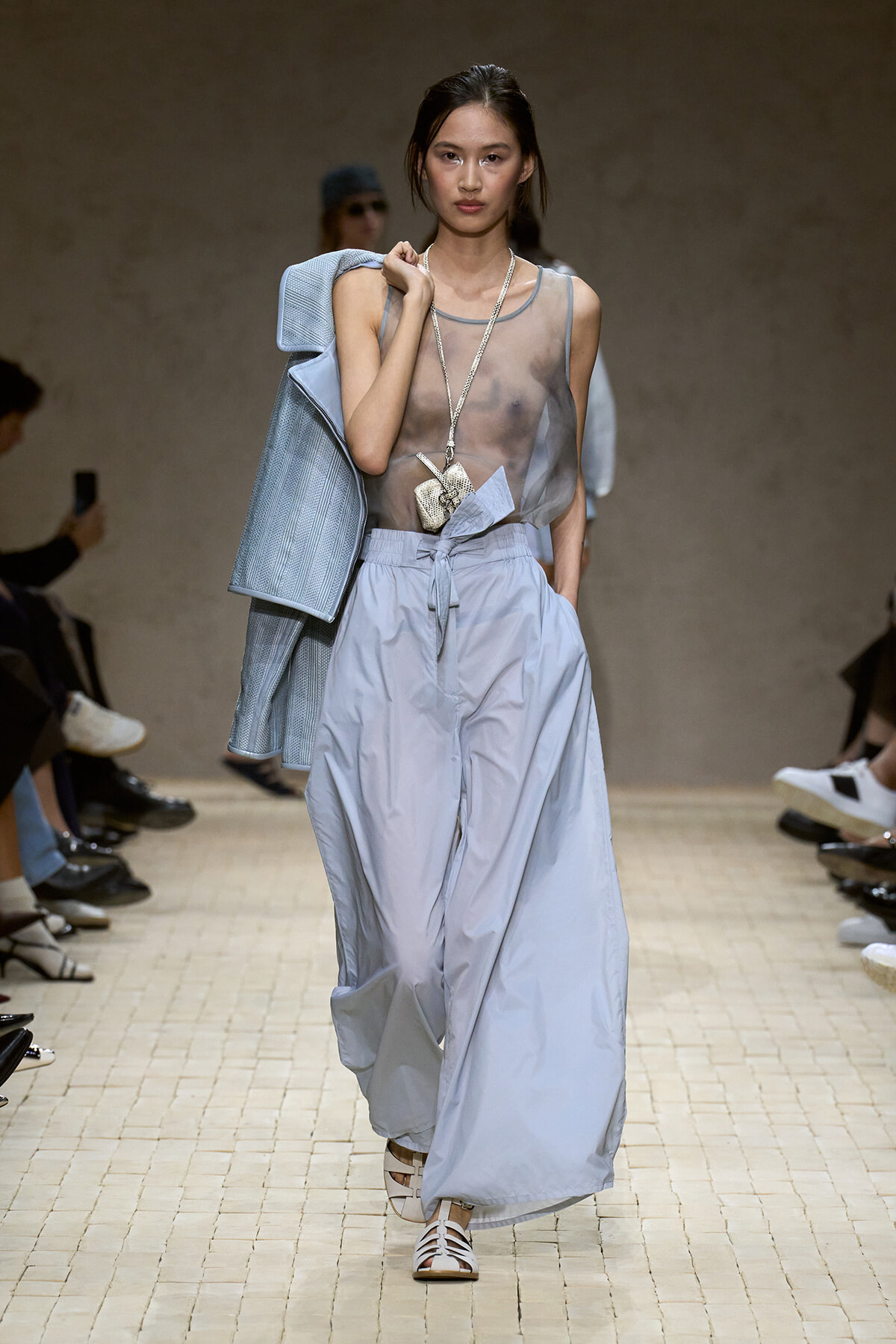 Model walks a runway in a sheer gray sleeveless top, pale blue wide-leg pants, a blue jacket over her shoulder, and a long necklace with a small pouch.