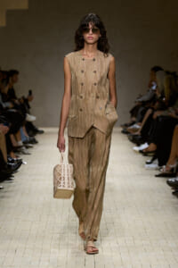 Model walking a runway in a beige sleeveless double‑breasted vest with matching wide‑leg pants, carrying a cream lattice handbag and wearing oversized round sunglasses.