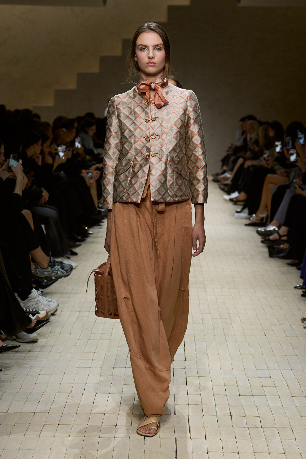 Model walks the runway in a silver quilted jacket with a peach scarf and matching peach wide-leg pants, carrying a brown lattice bag.