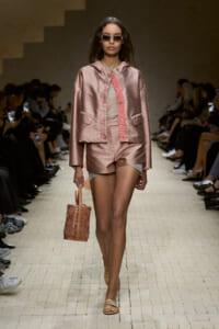 Runway model in a pink metallic jacket and matching shorts, carrying a woven pink handbag, walking the catwalk.