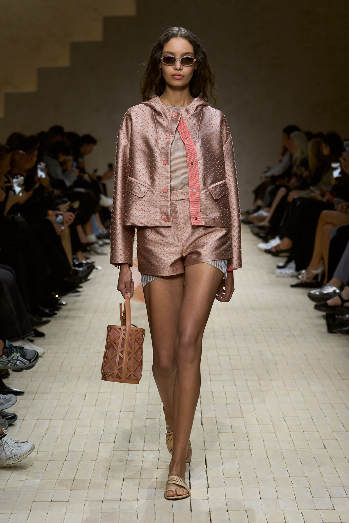 Runway model in a pink metallic jacket and matching shorts, carrying a woven pink handbag, walking the catwalk.