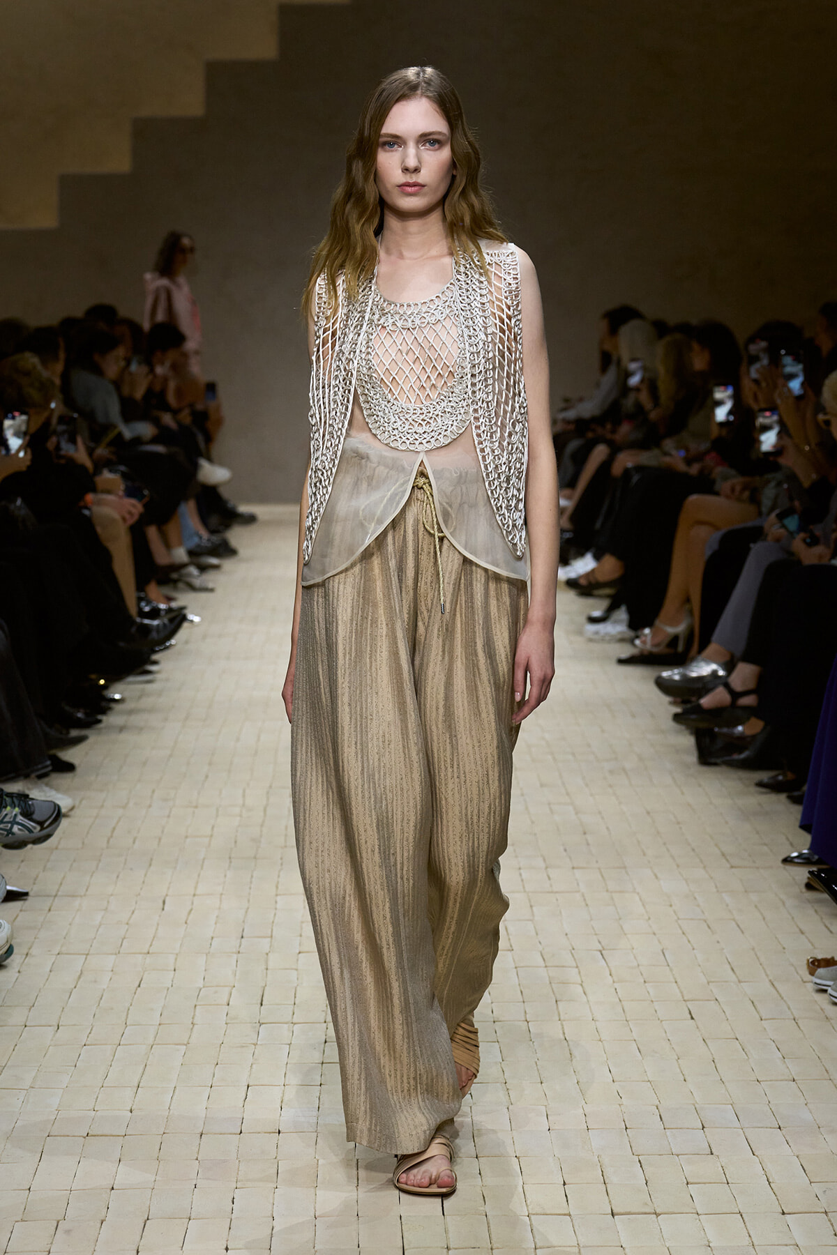 Model walking the runway in a sleeveless, silver mesh outer layer over a sheer top and wide-leg metallic beige pants; sandals and seated audience visible in background.
