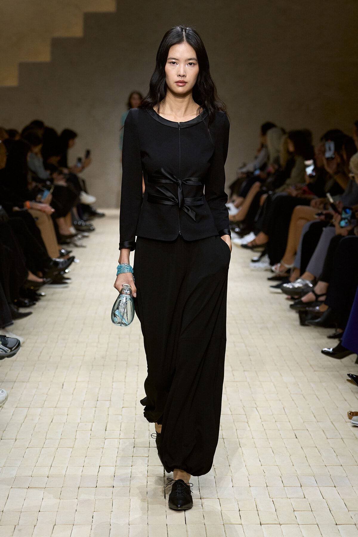 Model walking down a runway in a black tailored outfit with a wrapped belt, holding a small metallic bag and wearing a turquoise bracelet.