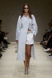 Model on a runway wearing a long light gray coat over white shorts, sunglasses, strappy white sandals, and a small snakeskin tote.