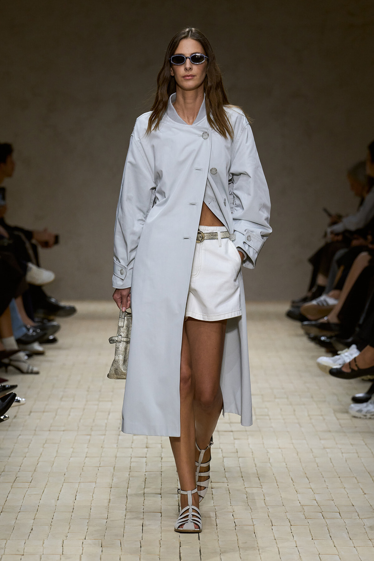 Model on a runway wearing a long light gray coat over white shorts, sunglasses, strappy white sandals, and a small snakeskin tote.