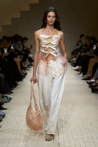 Model walking a runway in a sheer peach top with ribbon lacing and wide white pants, carrying a soft pink handbag.
