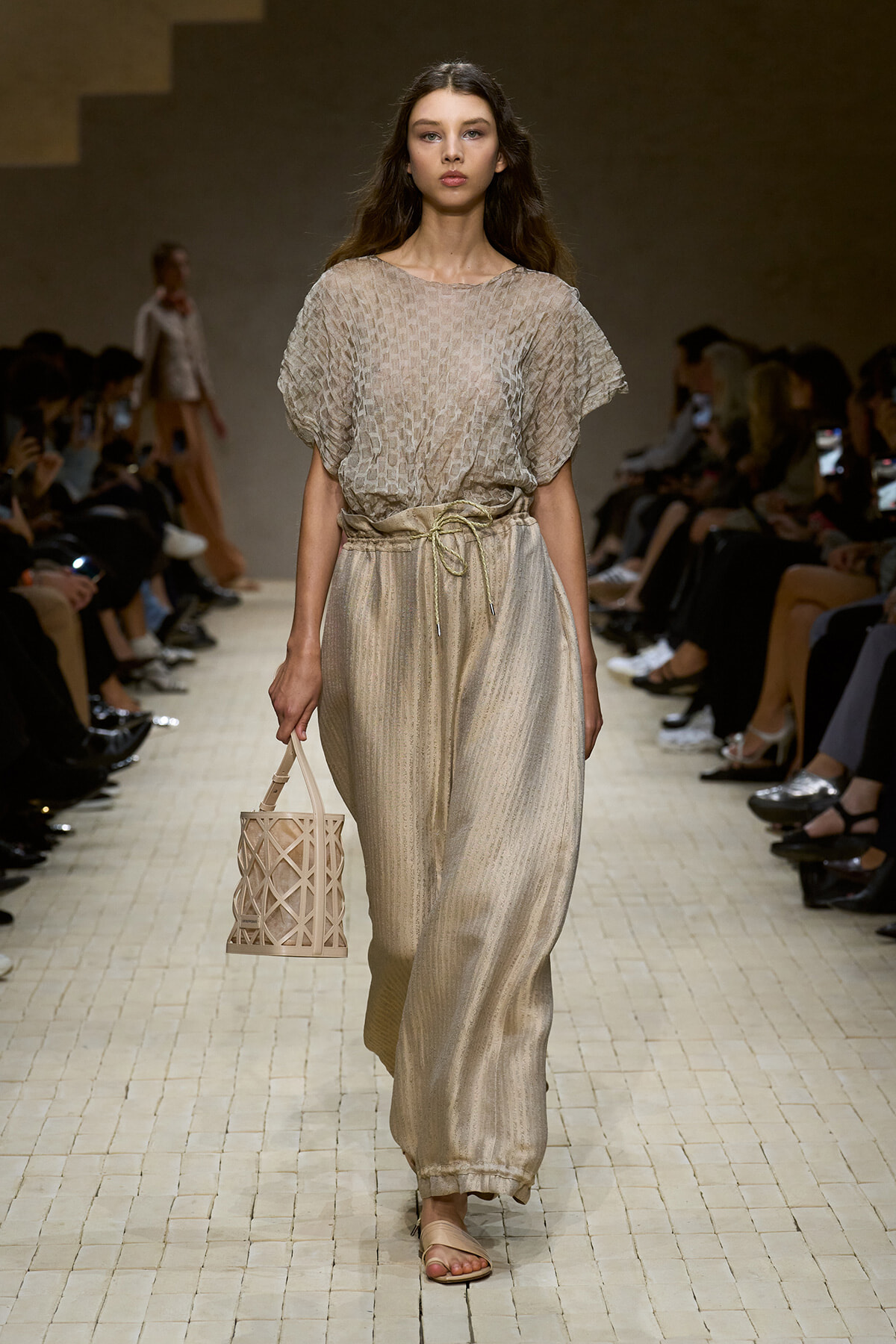Female model walking a runway in a beige textured blouse and wide-leg pants, carrying a lattice cream handbag and wearing flat sandals.