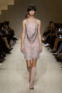 Model walking a runway in a pale pink beaded dress with draped chains and knee-high pink boots, audience watching on both sides.