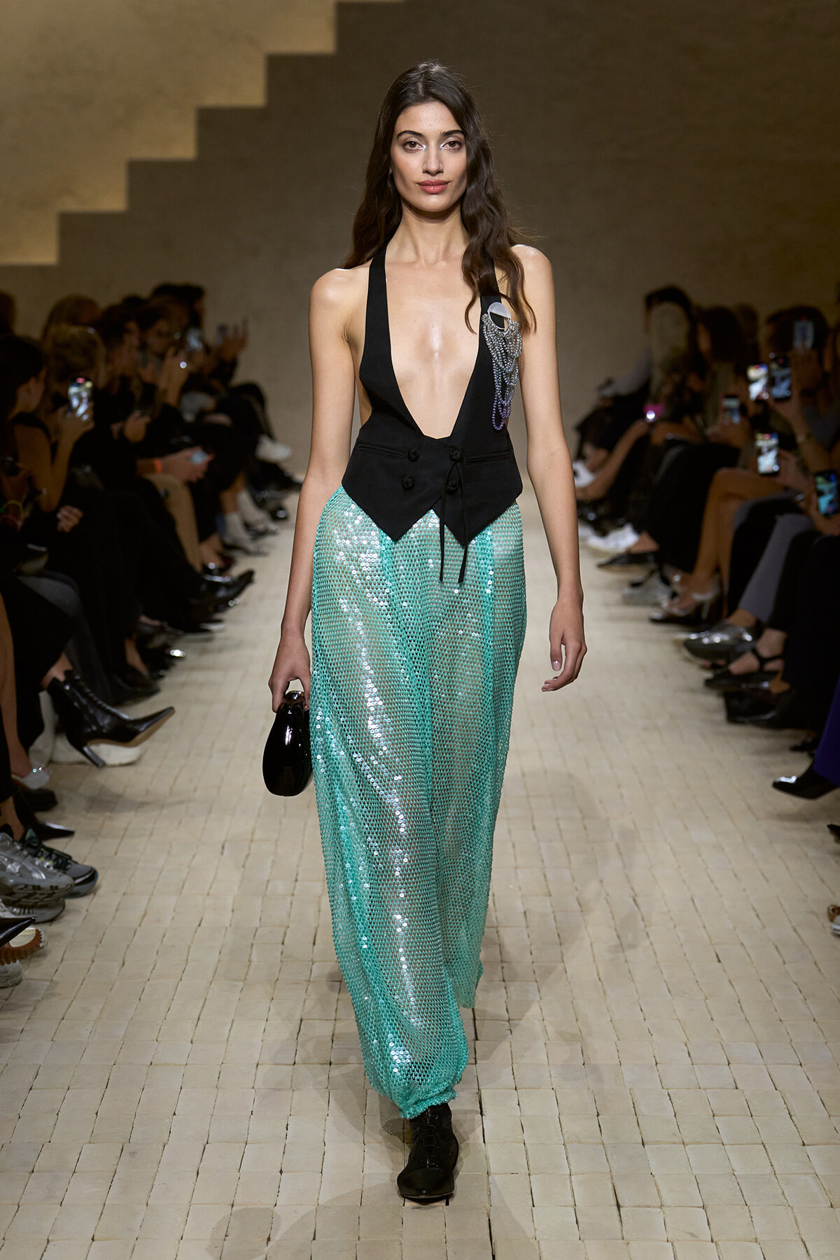Model walks a runway in a black plunging halter vest and turquoise sequin pants, holding a small black clutch.