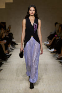 Female model walks a runway in a sleeveless black vest with purple bead brooch over lilac sequin wide-leg pants; carries a small black clutch.