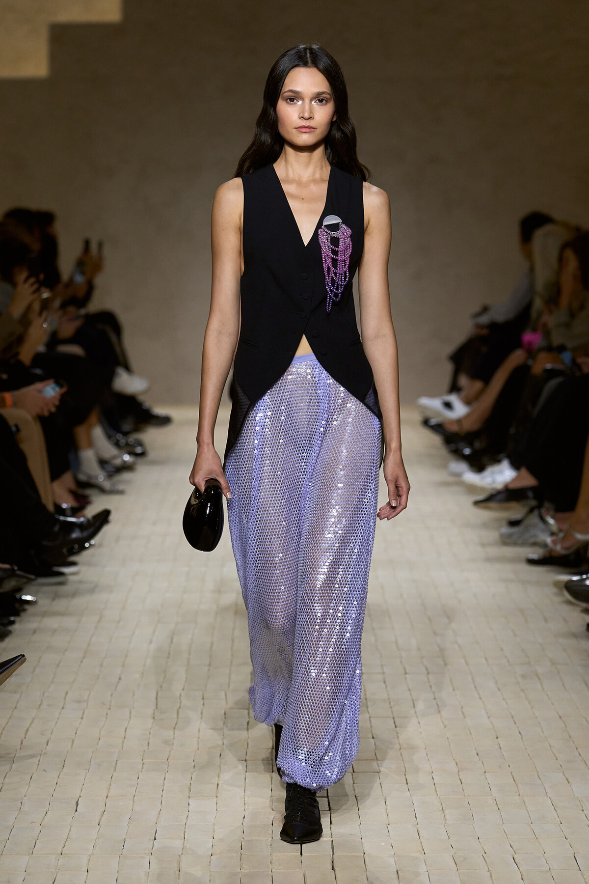 Female model walks a runway in a sleeveless black vest with purple bead brooch over lilac sequin wide-leg pants; carries a small black clutch.