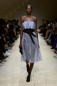 Model walking a fashion runway in a strapless light-blue dress with a wide black sash belt, black lace-up boots, and a small clutch; audience seated on both sides.