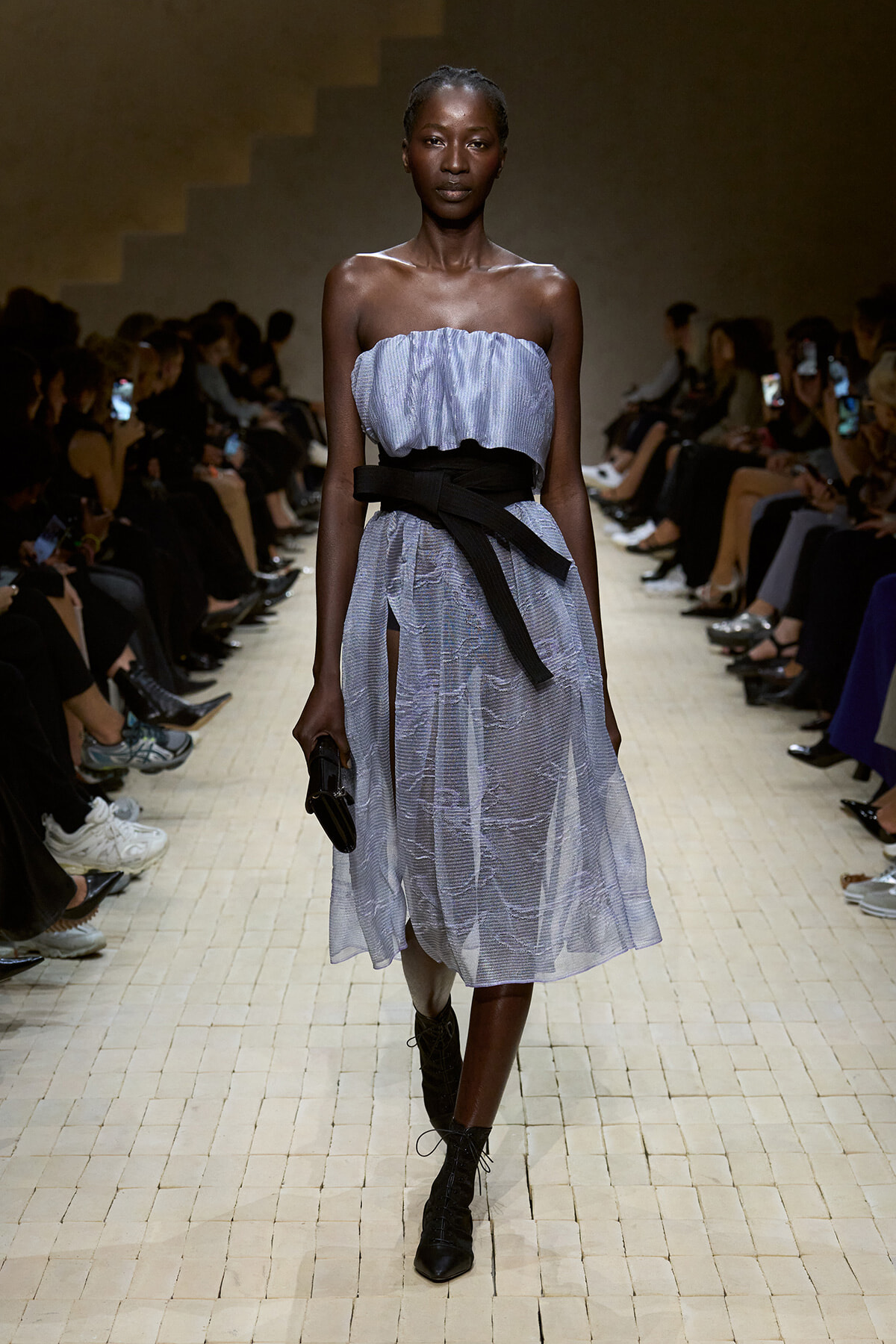 Model walking a fashion runway in a strapless light-blue dress with a wide black sash belt, black lace-up boots, and a small clutch; audience seated on both sides.