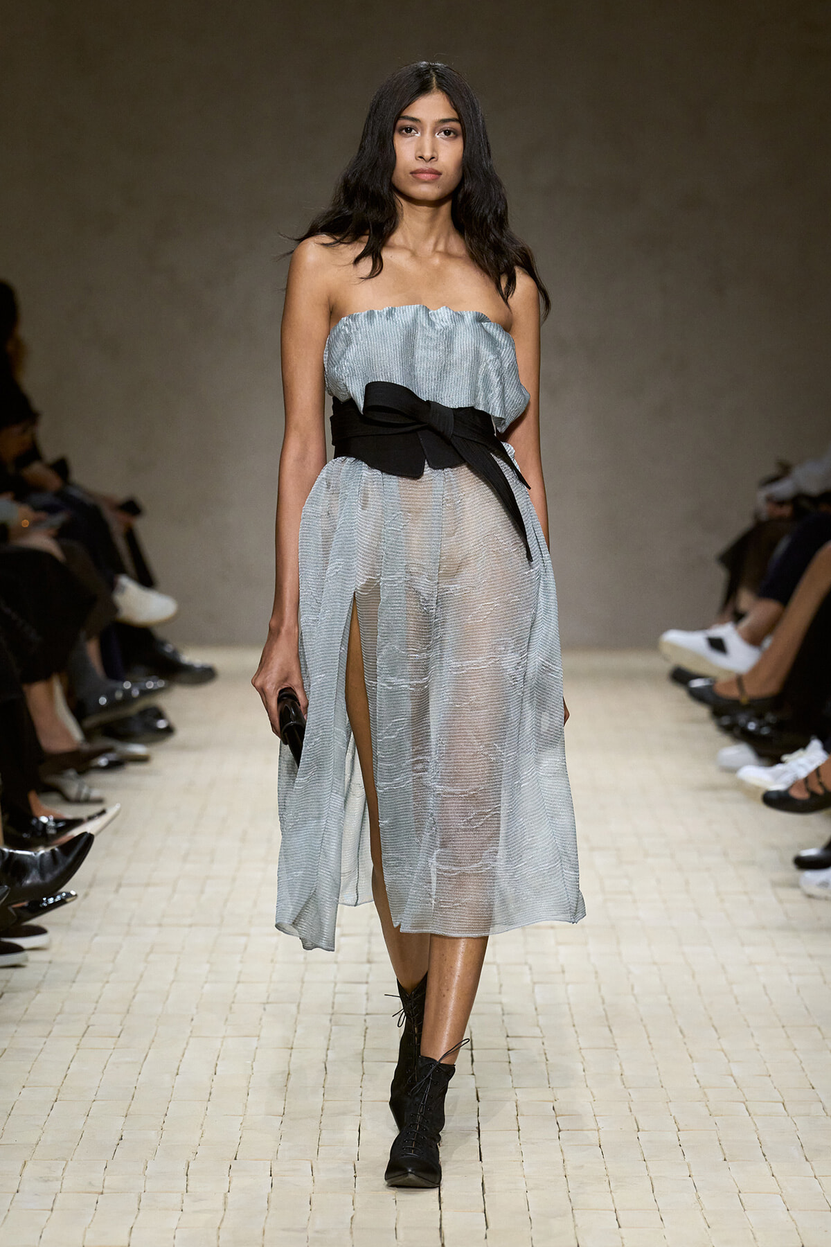 Model walking the runway in a light-blue strapless sheer midi dress with a thick black belt and lace-up black boots.