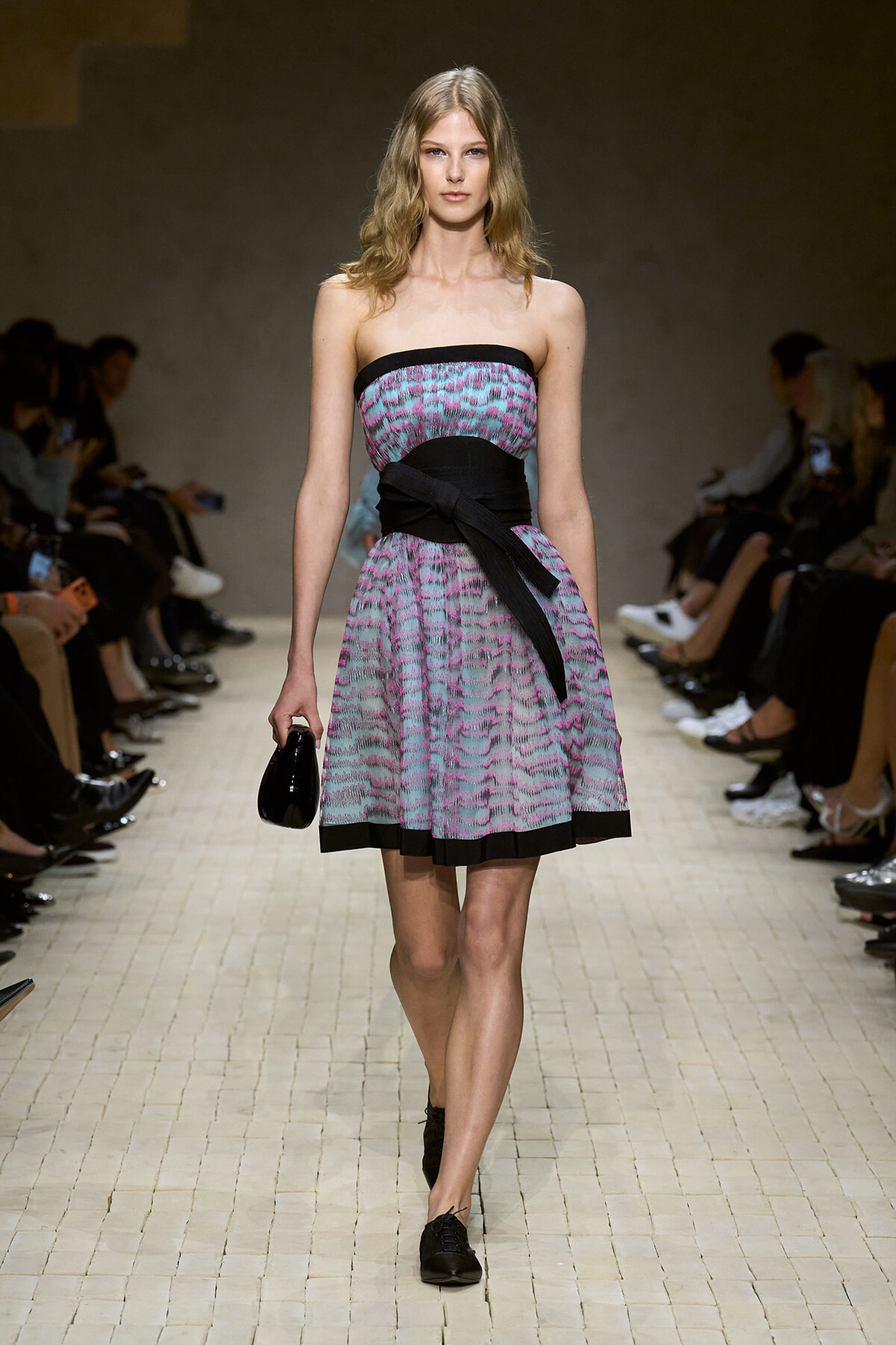 Model walks a fashion runway wearing a strapless patterned dress with a black waist sash, carrying a small black clutch as spectators watch from the sides.