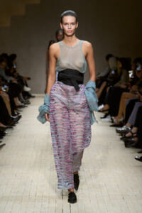 Model walking a runway in a silver mesh sleeveless top with a wide black belt, pink and gray patterned wide-leg pants, and light blue knit arm wraps, audience in background.
