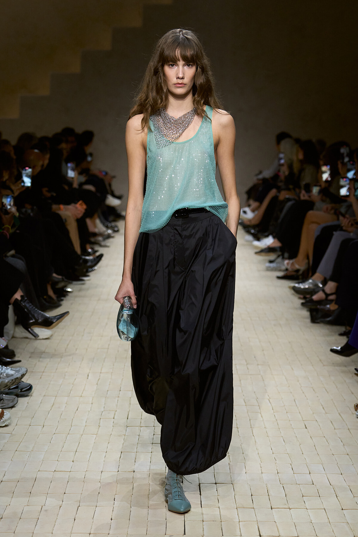 Model walks a fashion show wearing a mint-green sequined tank and oversized black balloon pants, turquoise shoes, and a small clear purse.