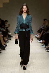 Model walking a runway in a teal patterned top with teal trim, a wide black belt, and loose black pants, clutch in hand.