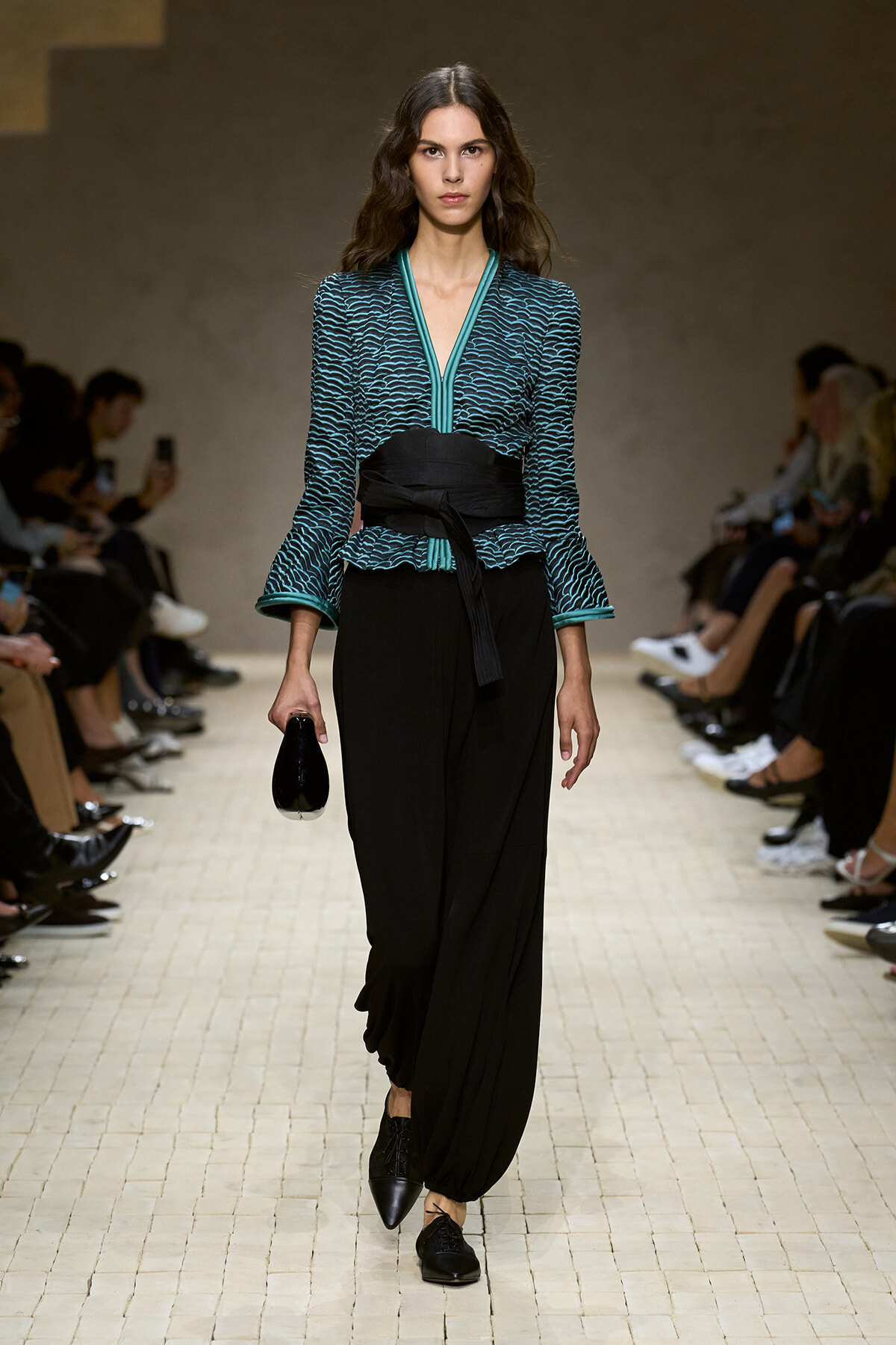 Model walking a runway in a teal patterned top with teal trim, a wide black belt, and loose black pants, clutch in hand.