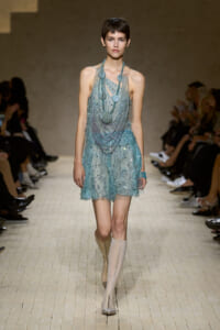 Model walking a fashion runway in a turquoise beaded mini dress with layered necklaces and tall boots.