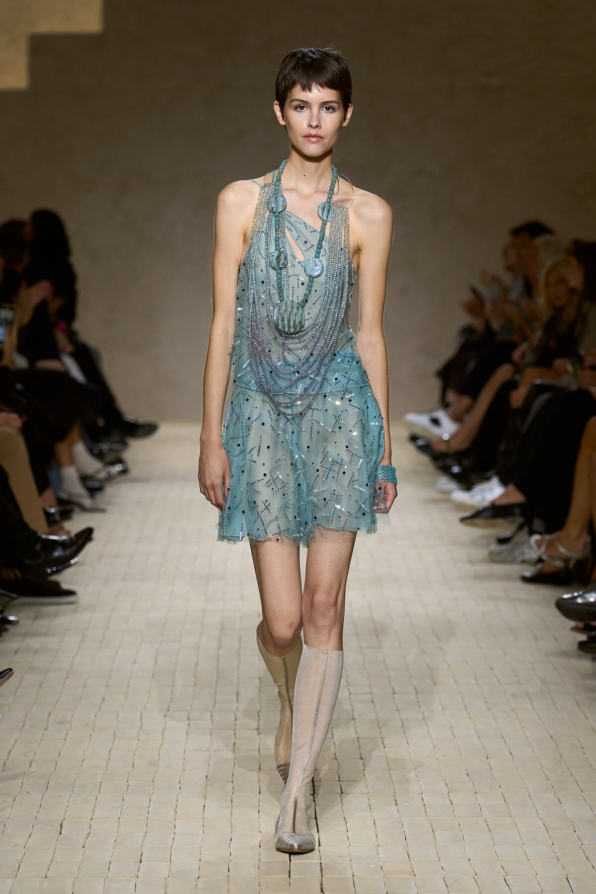 Model walking a fashion runway in a turquoise beaded mini dress with layered necklaces and tall boots.