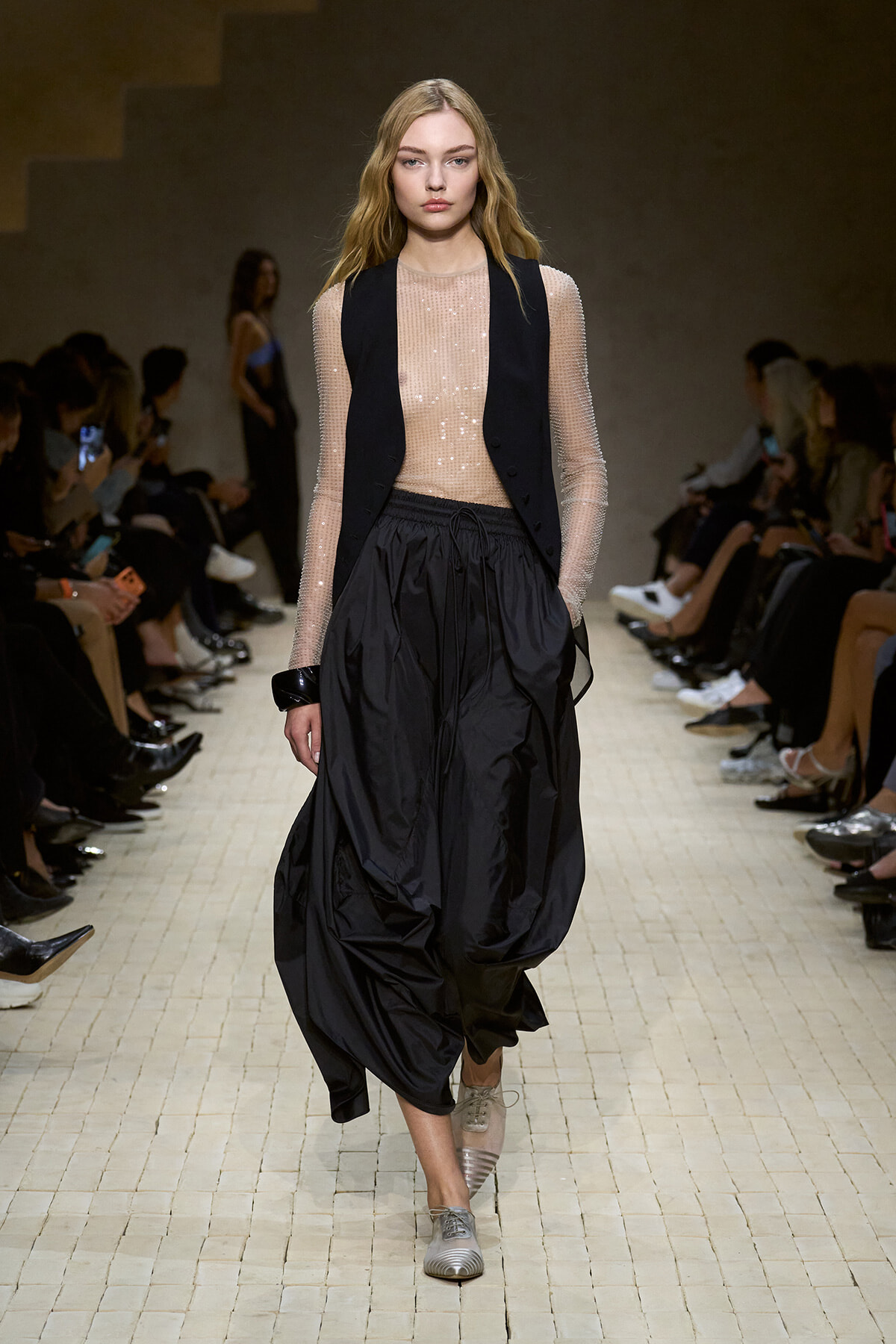 Model walks the runway in a sheer beaded top, black vest, and a voluminous black skirt, paired with metallic oxford shoes.