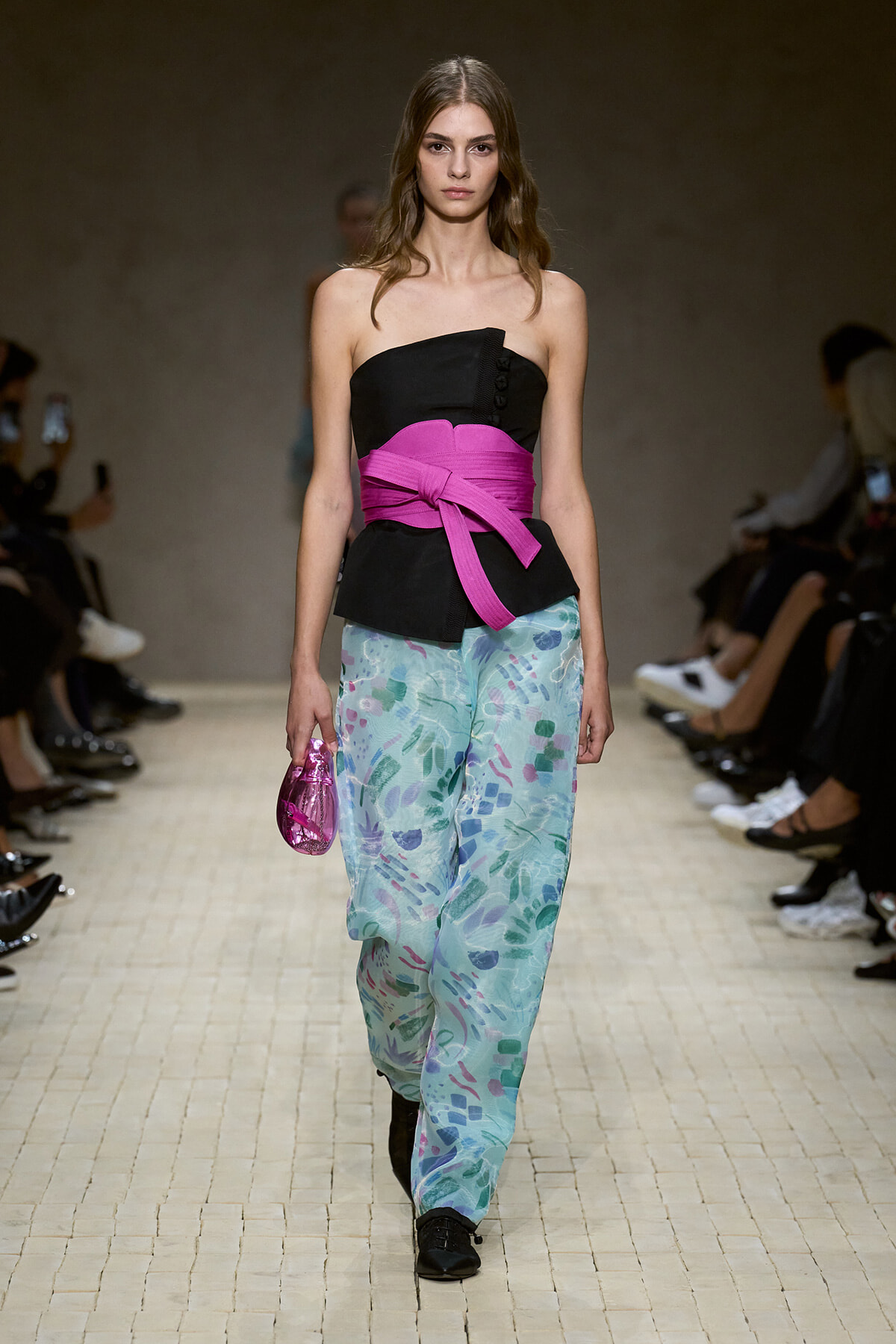 Model walks the runway in a black strapless top with a bright pink sash, turquoise floral-patterned pants, and a pink clutch.