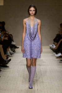 Model walks the runway in a lavender bead-embellished mini dress with layered silver chains and matching lilac boots, at a fashion show.