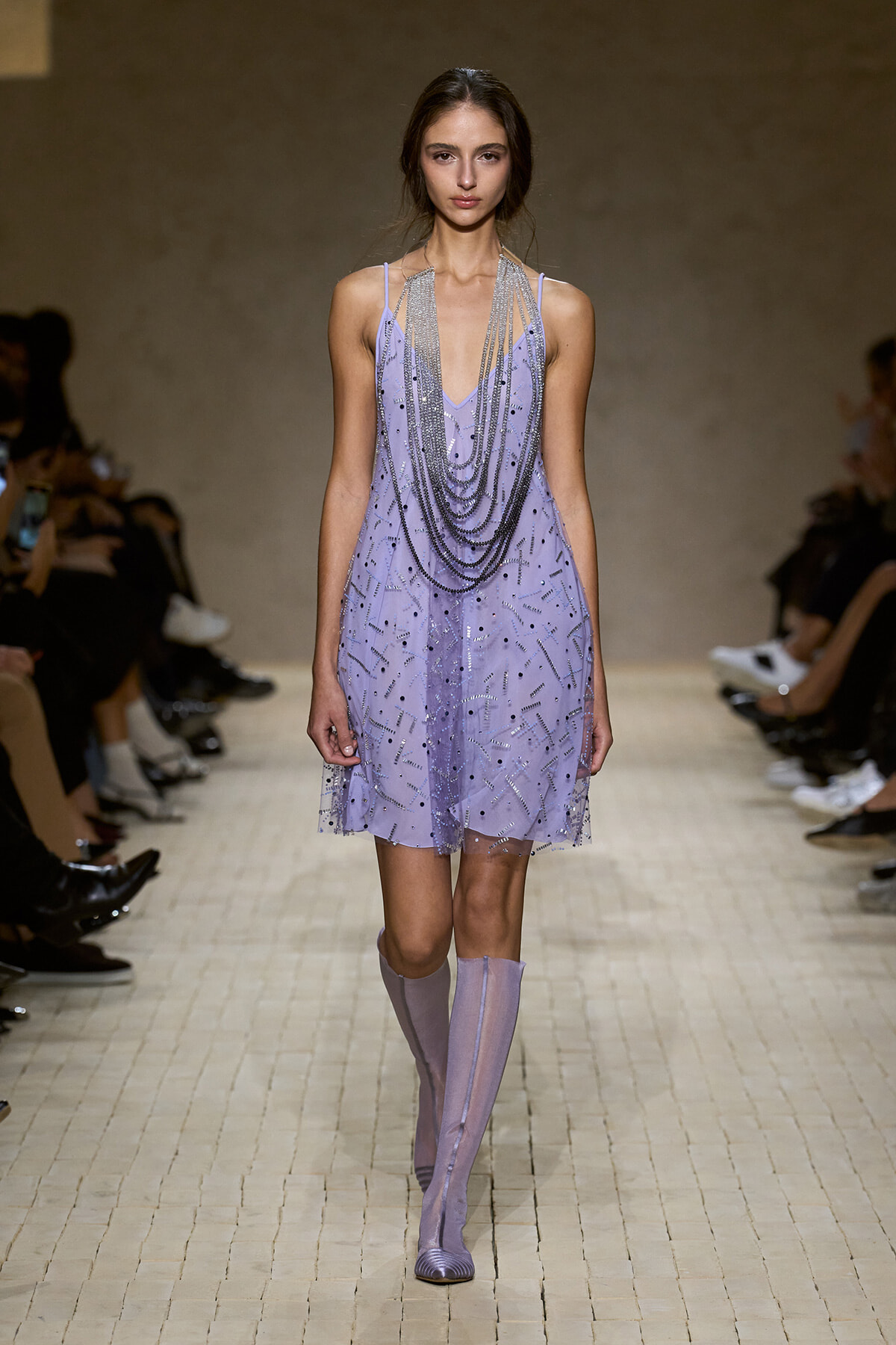Model walks the runway in a lavender bead-embellished mini dress with layered silver chains and matching lilac boots, at a fashion show.