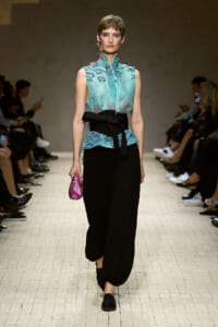 Model on a runway wearing a turquoise sleeveless embroidered top with a black waist belt and black wide pants, carrying a pink clutch.