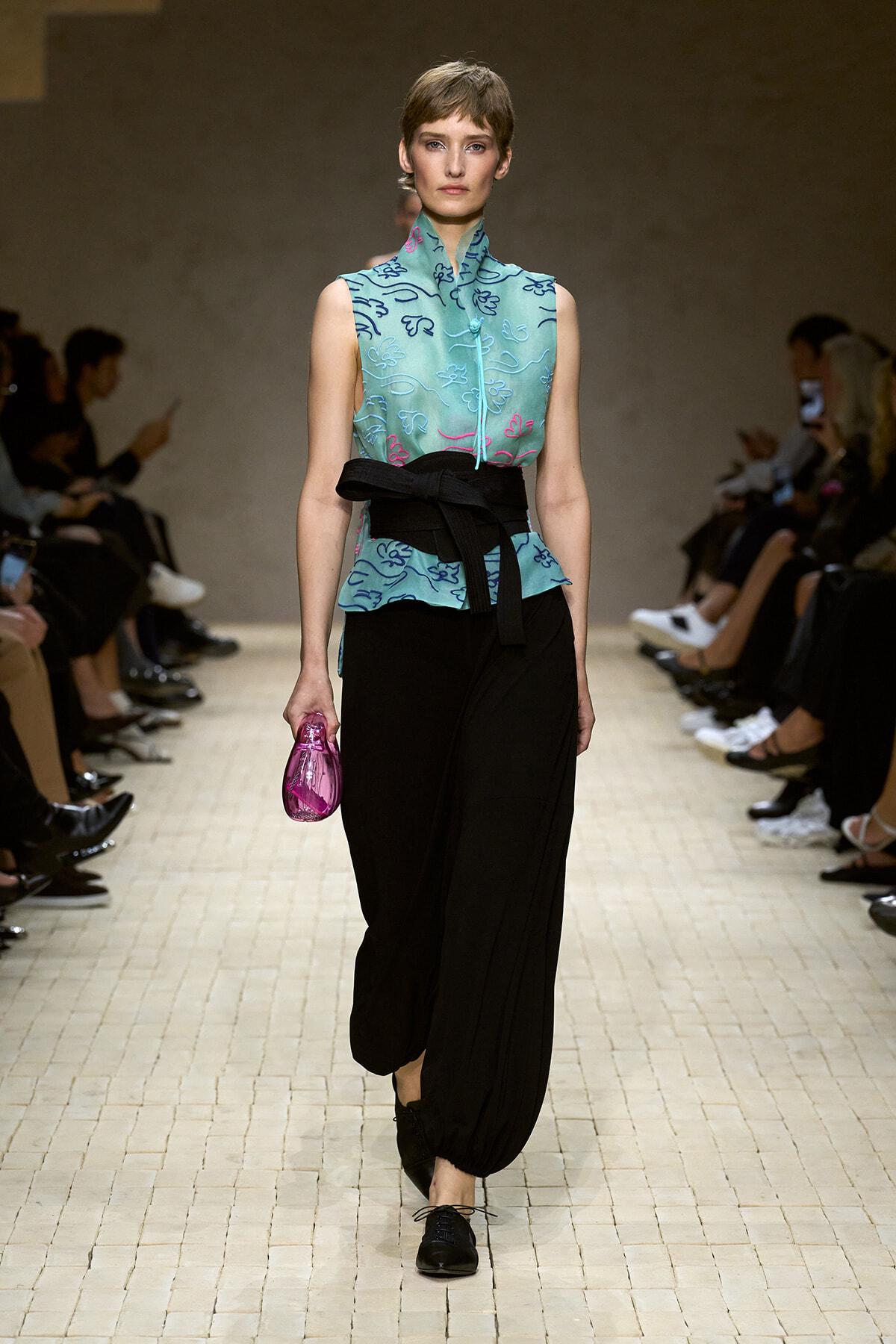 Model on a runway wearing a turquoise sleeveless embroidered top with a black waist belt and black wide pants, carrying a pink clutch.