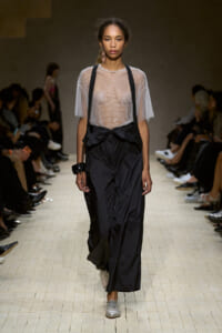 Model walks the runway in a sheer, beaded top paired with black, oversized pants with suspenders and silver shoes.