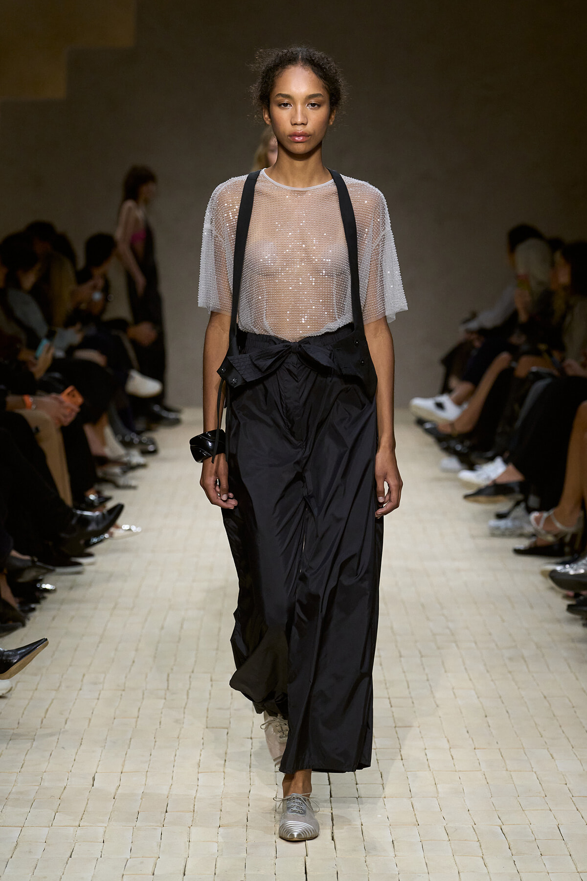 Model walks the runway in a sheer, beaded top paired with black, oversized pants with suspenders and silver shoes.