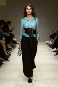 Model walks a runway in a teal embroidered blouse with a black obi belt and loose black pants, holding a small blue clutch.
