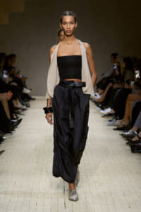 Model walking the runway in a black strapless top, cream draped vest, and oversized black cargos cinched with a belt.