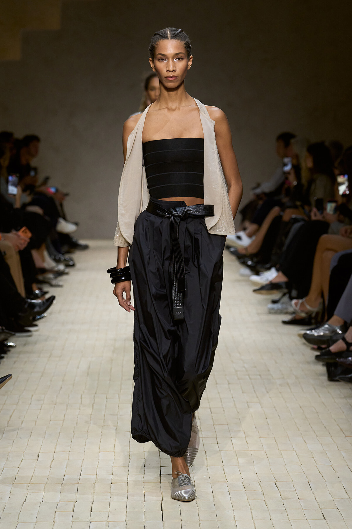 Model walking the runway in a black strapless top, cream draped vest, and oversized black cargos cinched with a belt.