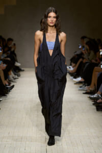 Model walks the runway in a black, oversized jumpsuit with a blue sequined bandeau top, audience seated along the catwalk behind.