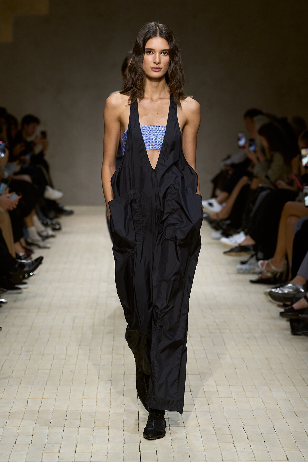 Model walks the runway in a black, oversized jumpsuit with a blue sequined bandeau top, audience seated along the catwalk behind.