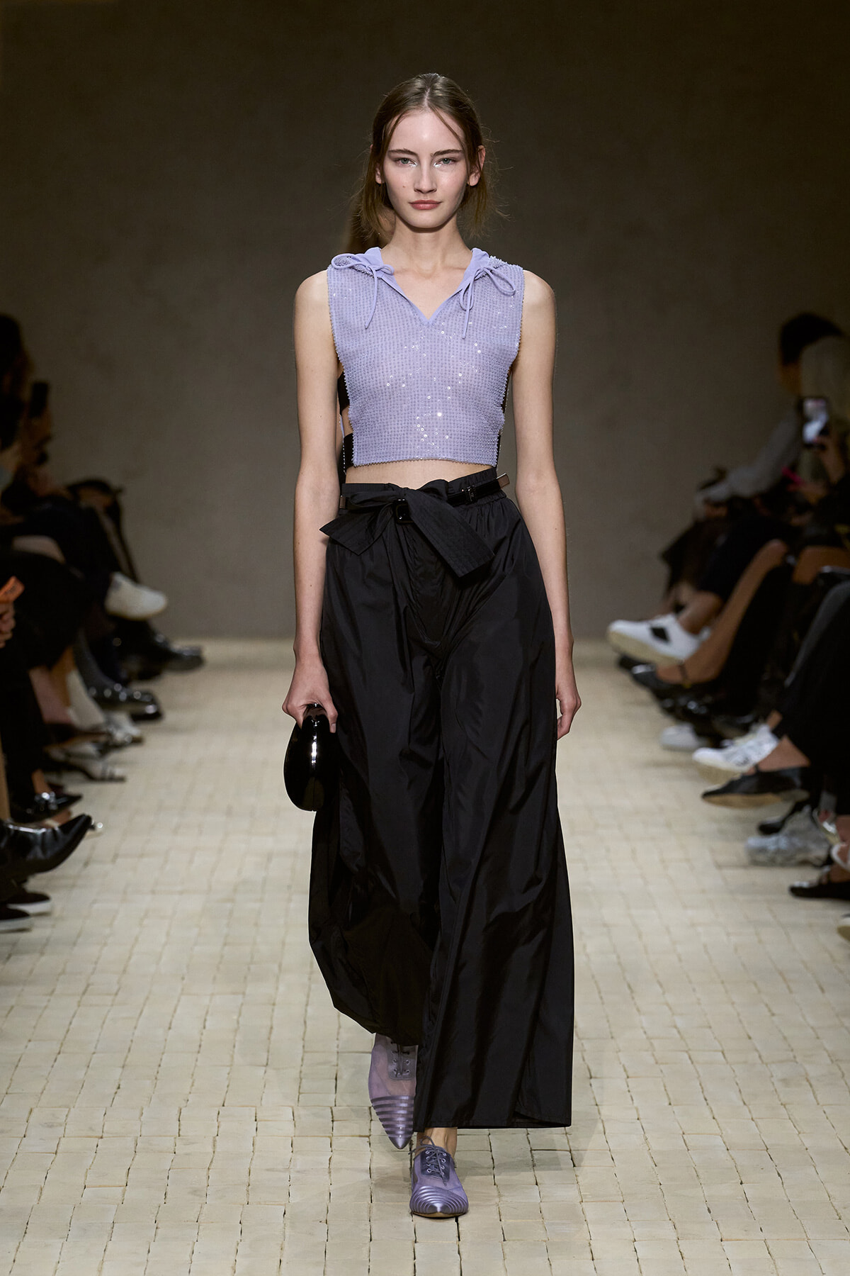 Model walking a fashion show in a lavender sequined cropped sleeveless hoodie with tie details, paired with high-waisted black wide-leg pants and a small black bag on her side.