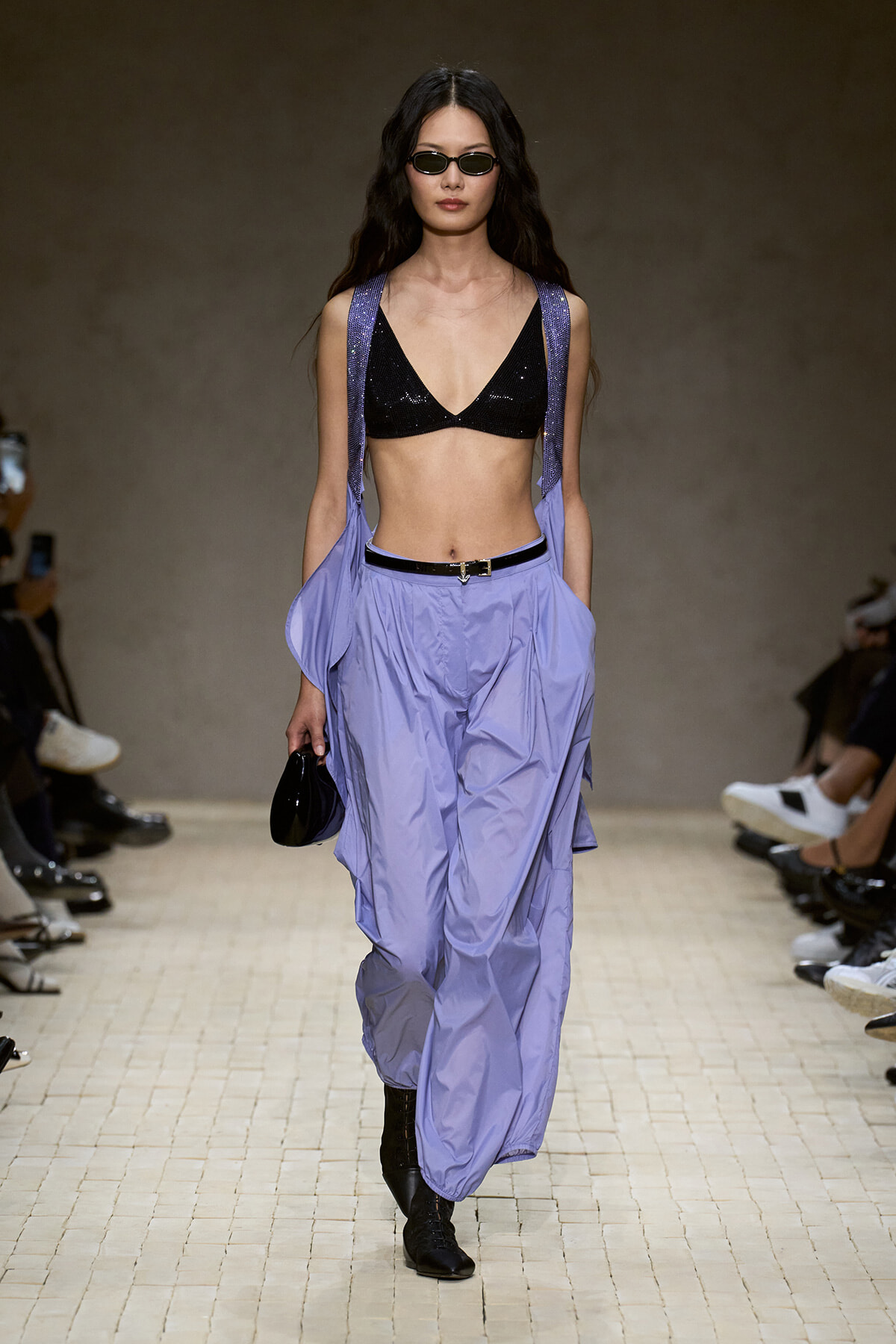 Model struts the runway in a black sequin bralette with lavender exaggerated-pocket pants and suspender straps, accessorized with a black belt, sunglasses, and a small black purse.